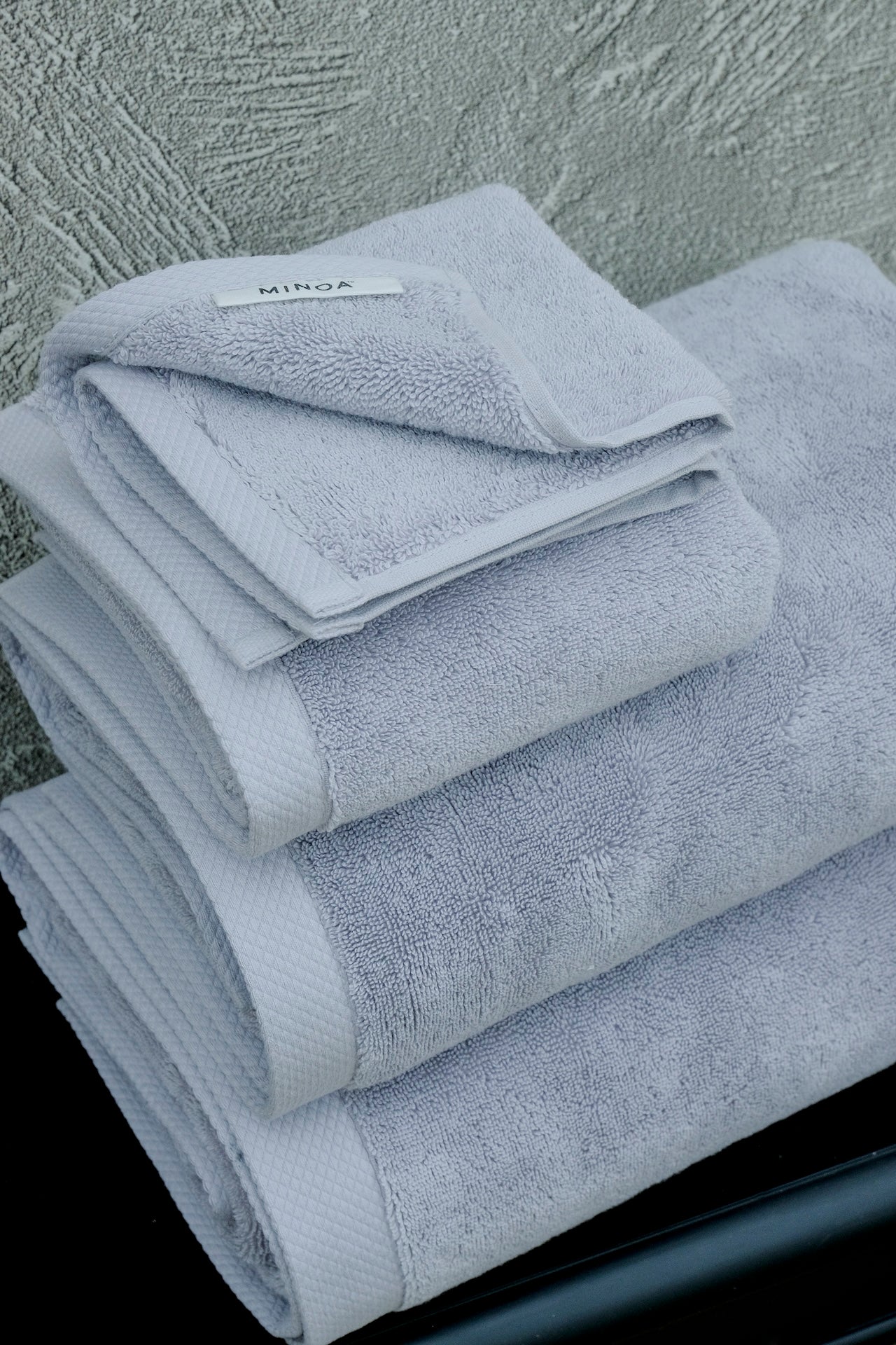 Stack of premium grey bath towels with visible MINOA tag, made from organic cotton, neatly folded against a textured wall.
