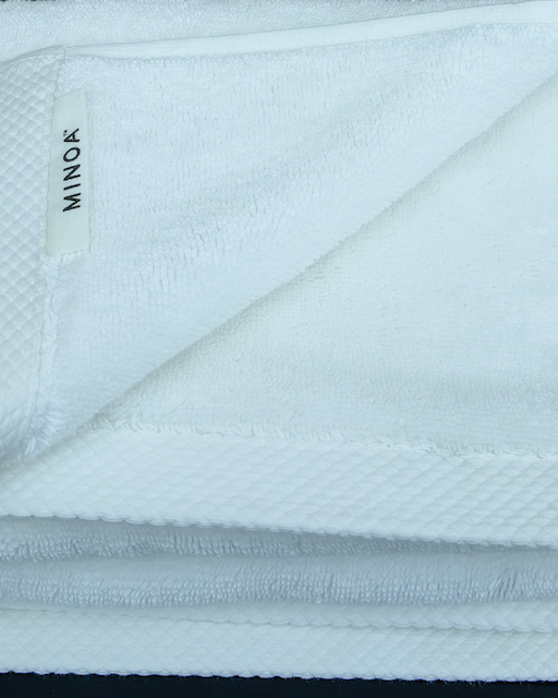 Close-up of a folded premium white bath towel with visible MINOA label, crafted from organic cotton and displayed on a dark surface.