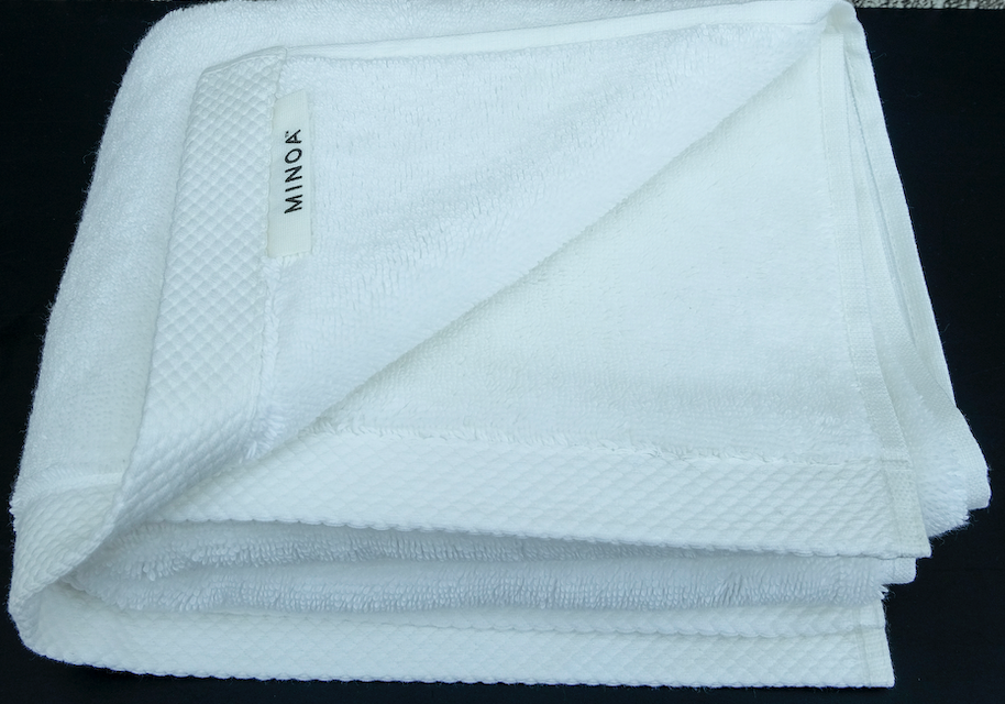 Close-up of a folded premium white bath towel with visible MINOA label, crafted from organic cotton and displayed on a dark surface.