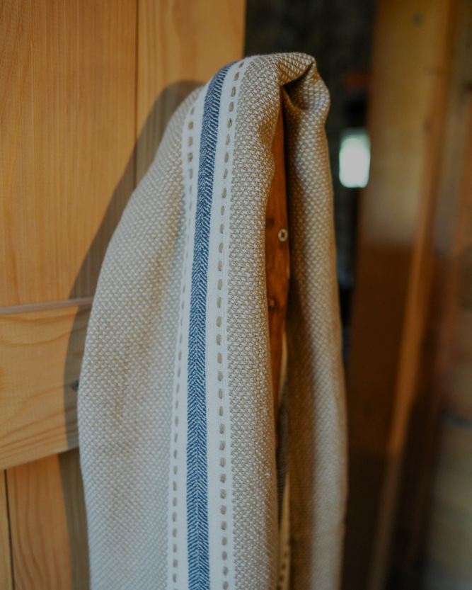 Handwoven sandstone Tulum 200 gram Aegean Cotton and Linen Turkish peshtemal towel by Minoa Home featuring beige and blue stripes hanging on a wooden hook showcasing the softness and quality of an organic bath towel