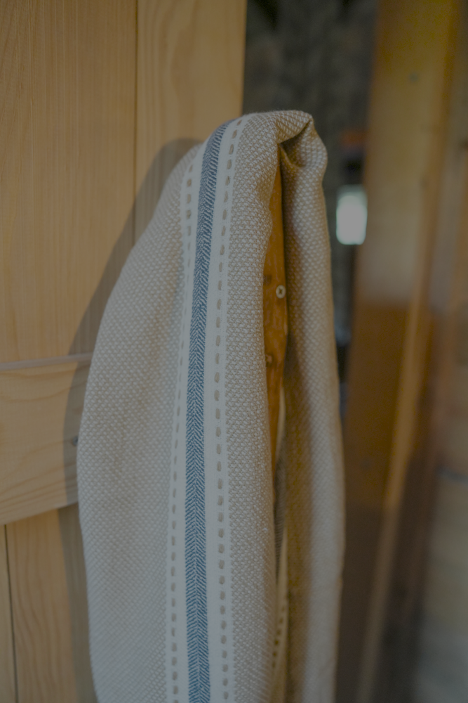 Handwoven sandstone Tulum 200 gram Aegean Cotton and Linen Turkish peshtemal towel by Minoa Home featuring beige and blue stripes hanging on a wooden hook showcasing the softness and quality of an organic bath towel