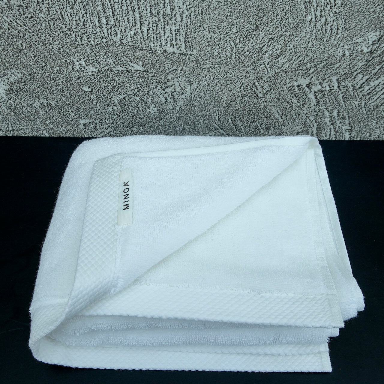 White luxury cotton towel neatly folded on a black surface with a textured grey wall background, showcasing softness and premium quality by Minoa Home.