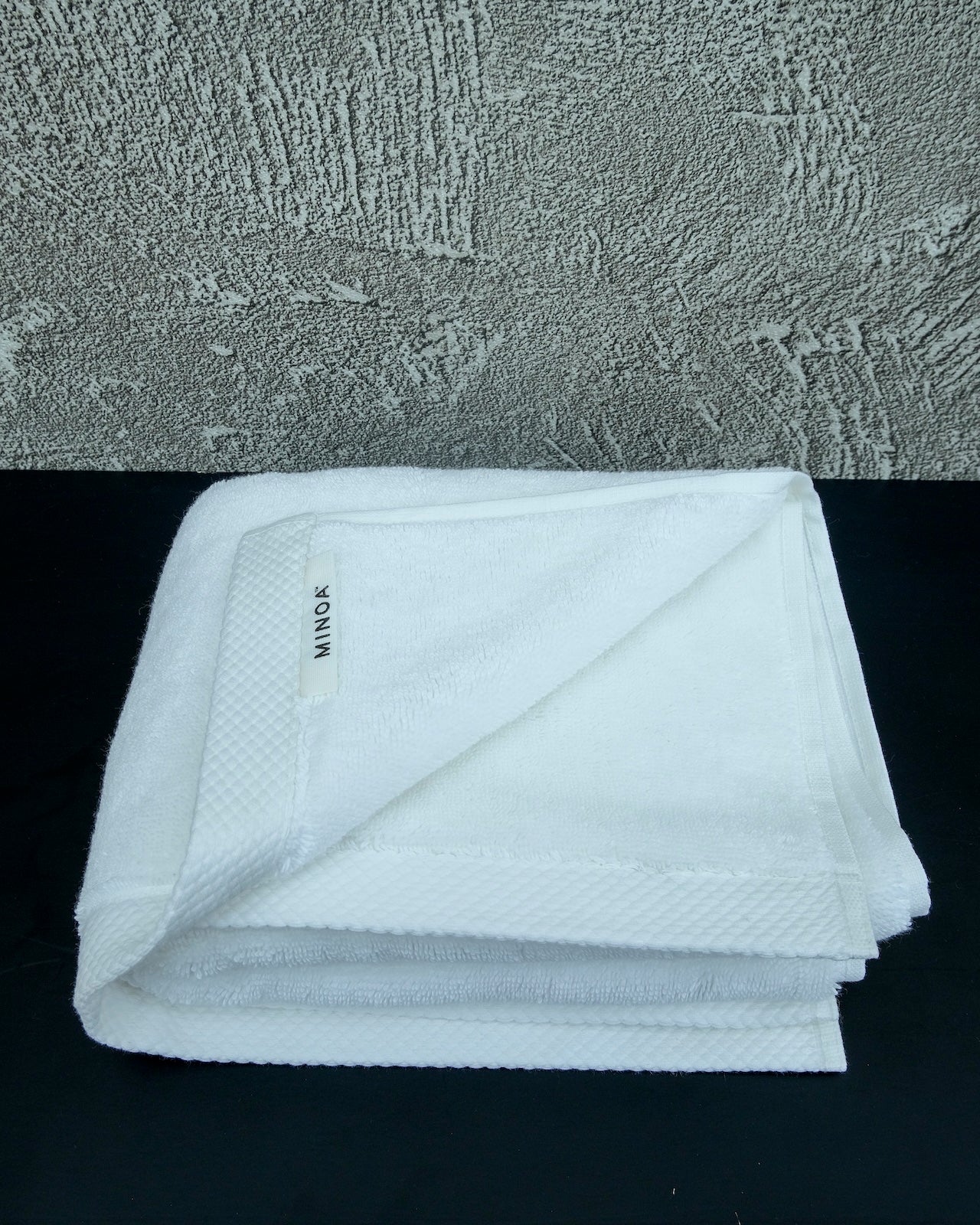 White luxury cotton towel neatly folded on a black surface with a textured grey wall background, showcasing softness and premium quality by Minoa Home.