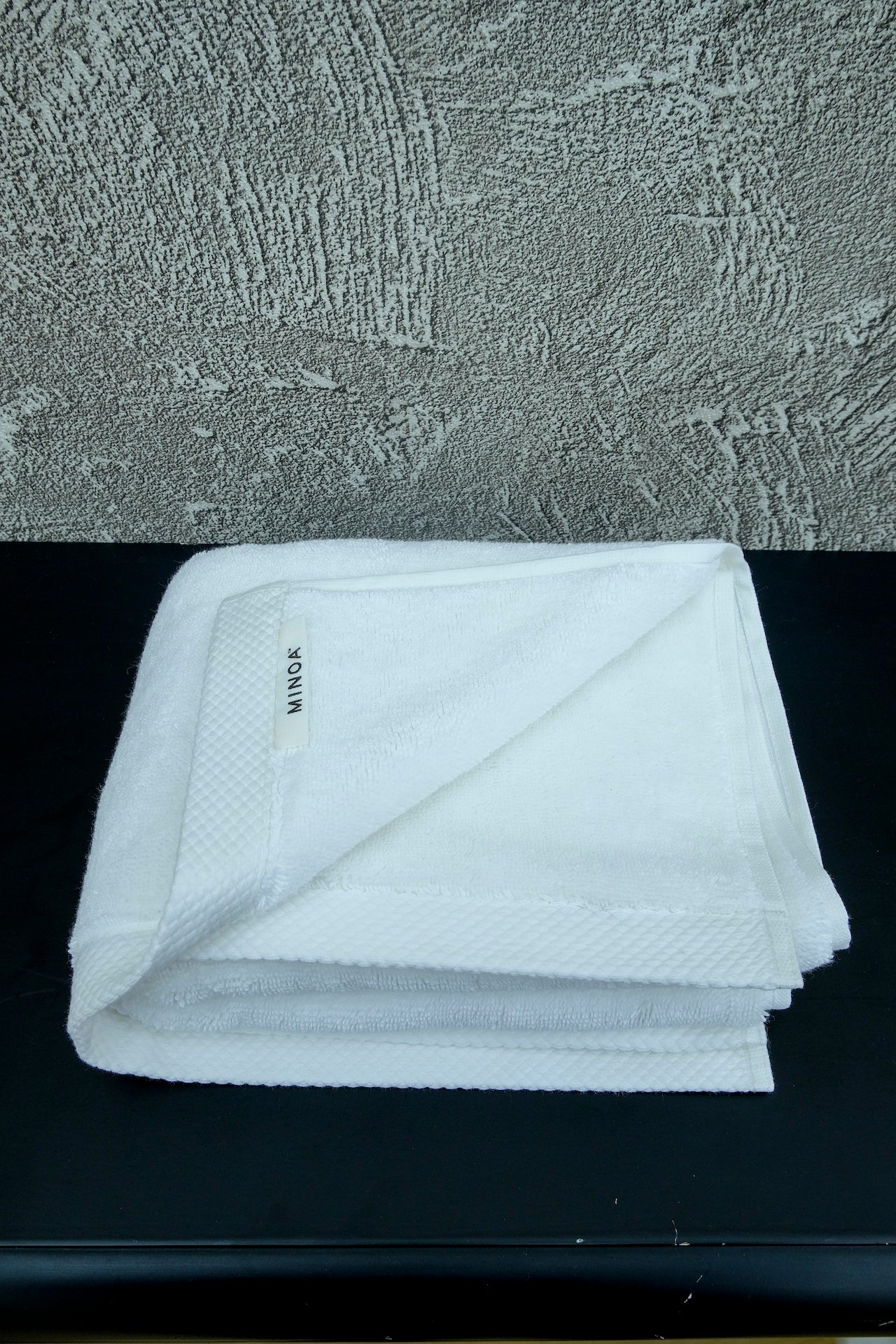 White luxury cotton towel neatly folded on a black surface with a textured grey wall background, showcasing softness and premium quality by Minoa Home.