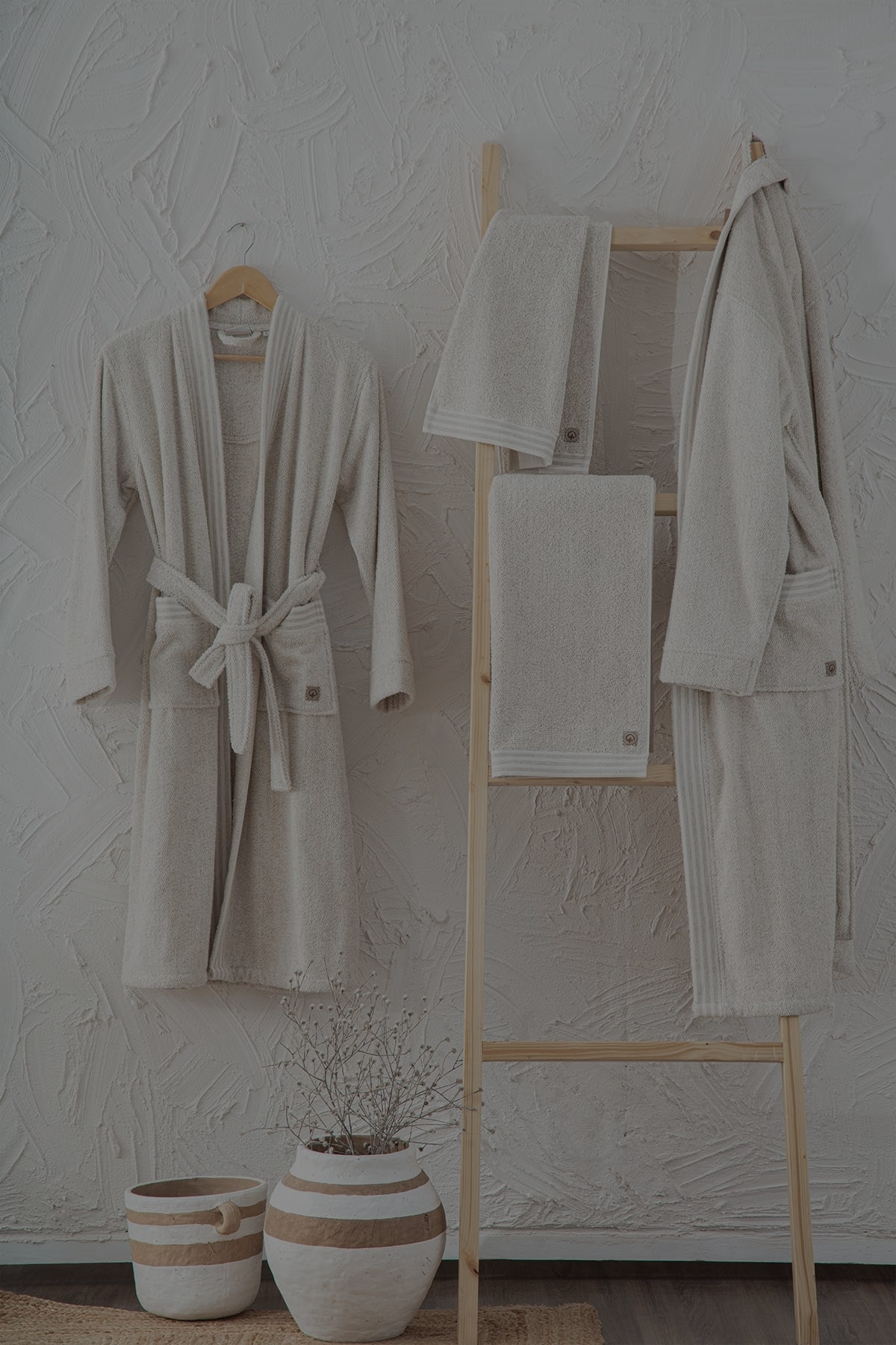 Neutral-toned luxury bathrobes and towels displayed on a wooden rack with matching slippers and storage baskets, showcasing Minoa Home's sustainable home essentials.