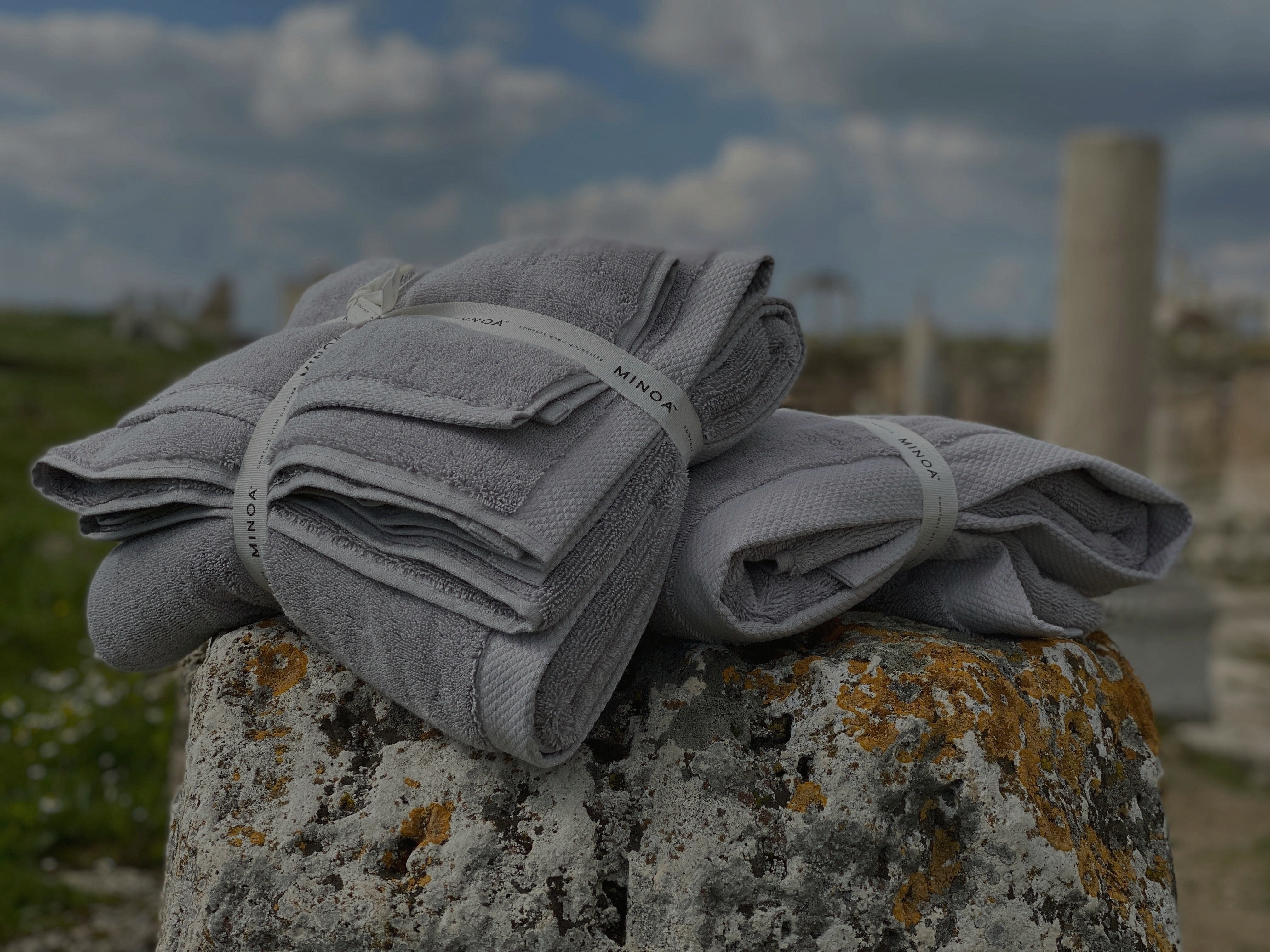 Stack of Minoa Home’s premium grey towels placed on ancient stone ruins under a blue sky, showcasing quality and durability