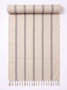 A beige woven towel with vertical dark gray stripes and fringed tassels along one edge, shown partially rolled on a plain white background.