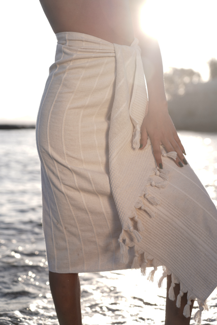 Person wearing a white towel with fringes by a body of water.