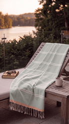 A pale mint green beach towel with white windowpane plaid lines and cream fringe, draped over a wooden lounge chair on a patio overlooking a lake at sunset.
