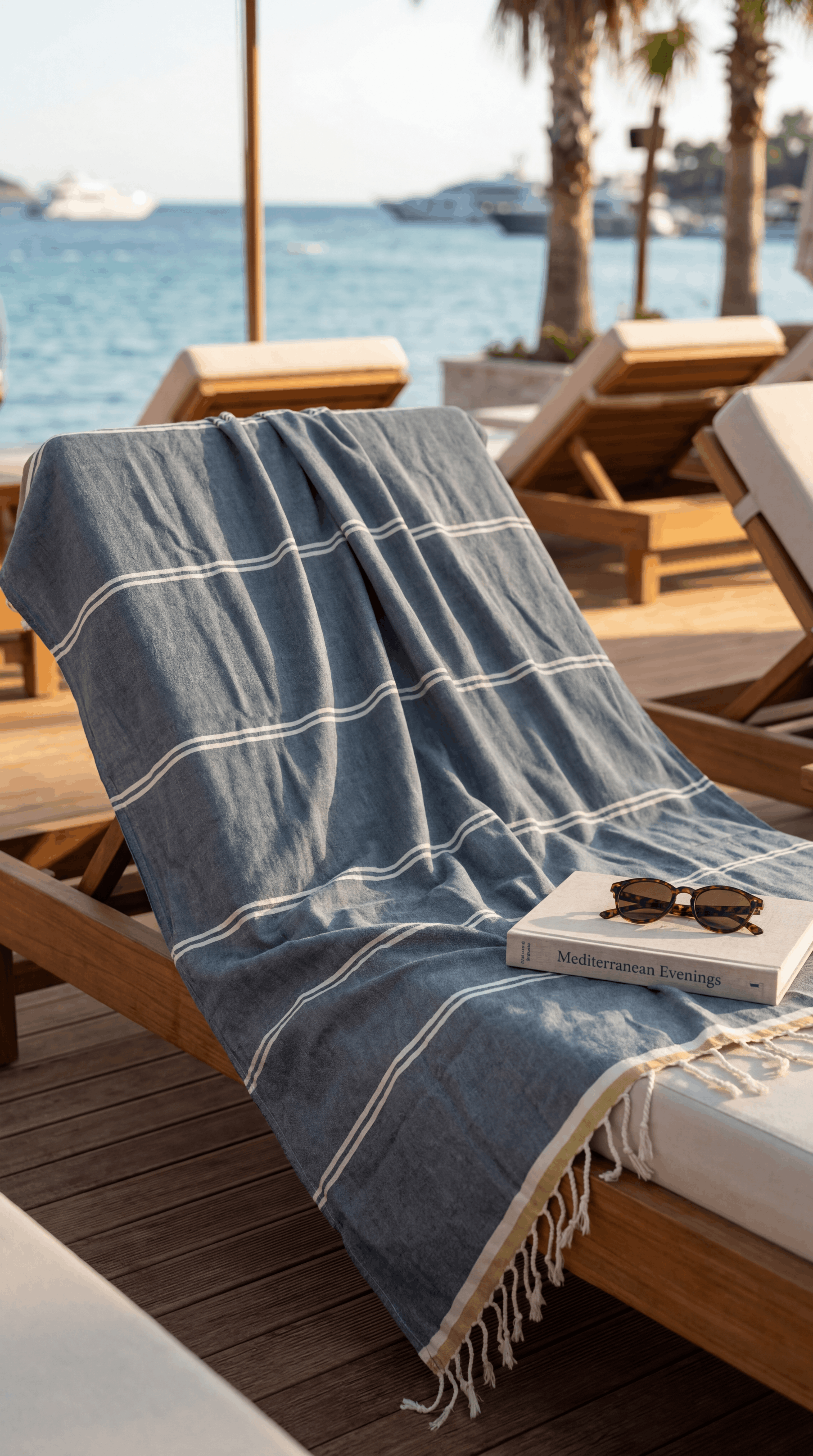 A blue and white striped cotton beach towel with fringe trim draped over a wooden lounge chair on a sunny beach deck, with the ocean and palm trees in the background.