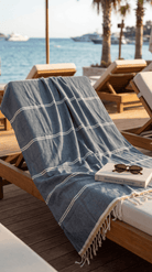 A blue and white striped cotton beach towel with fringe trim draped over a wooden lounge chair on a sunny beach deck, with the ocean and palm trees in the background.