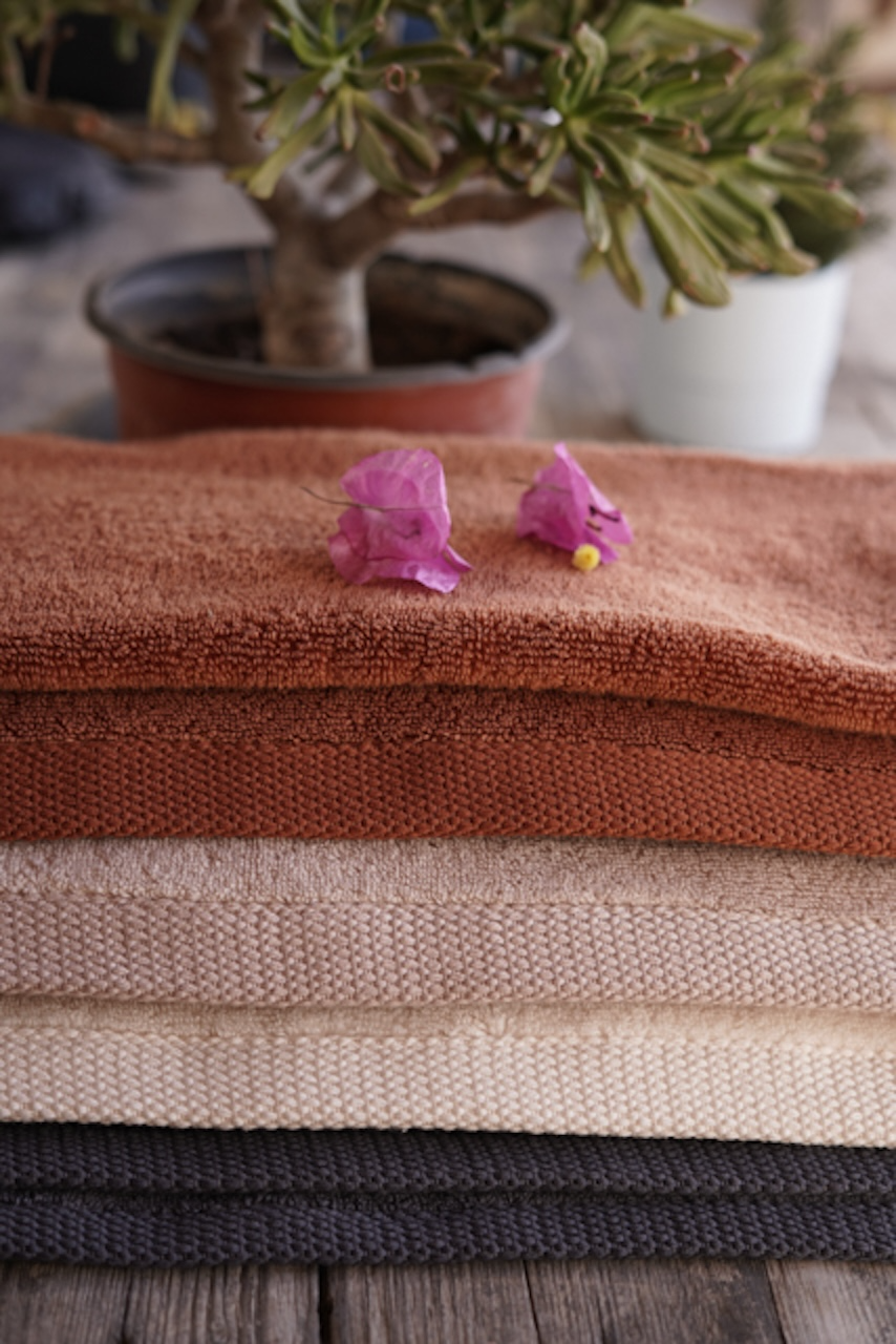 Plush Lite 500-Gram Aegean Cotton luxury bath towels in Cinnamon Charm, Honey Latte, Moonlight Pearl, and Charcoal Delight, neatly folded with a plant background, showcasing their soft texture and vibrant colours.

