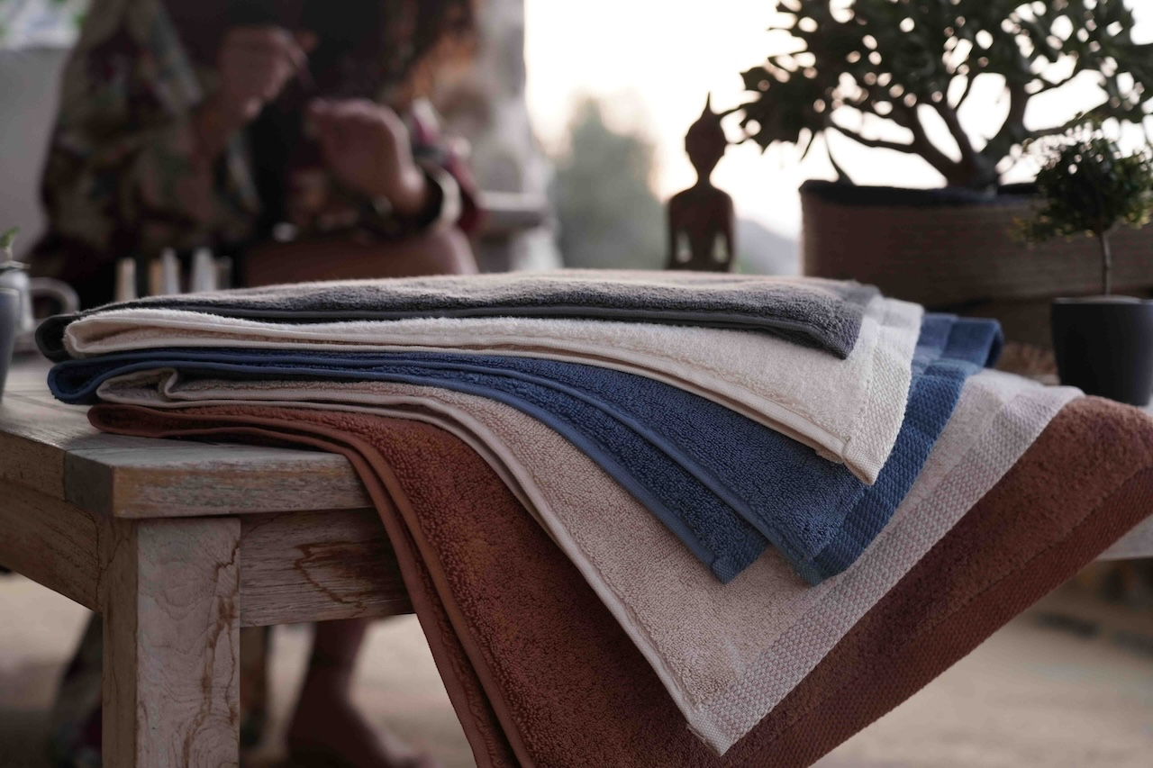 Plush Lite 500 gram Aegean Cotton large bath and hand towel set in Moonlight Pearl Honey Latte Charcoal Embrace Blue Velvet Cinnamon Charm and Pure n Raw arranged on a tree pattern table beside a woman showcasing sustainable luxury with soft organic bath towels and luxury bath towels