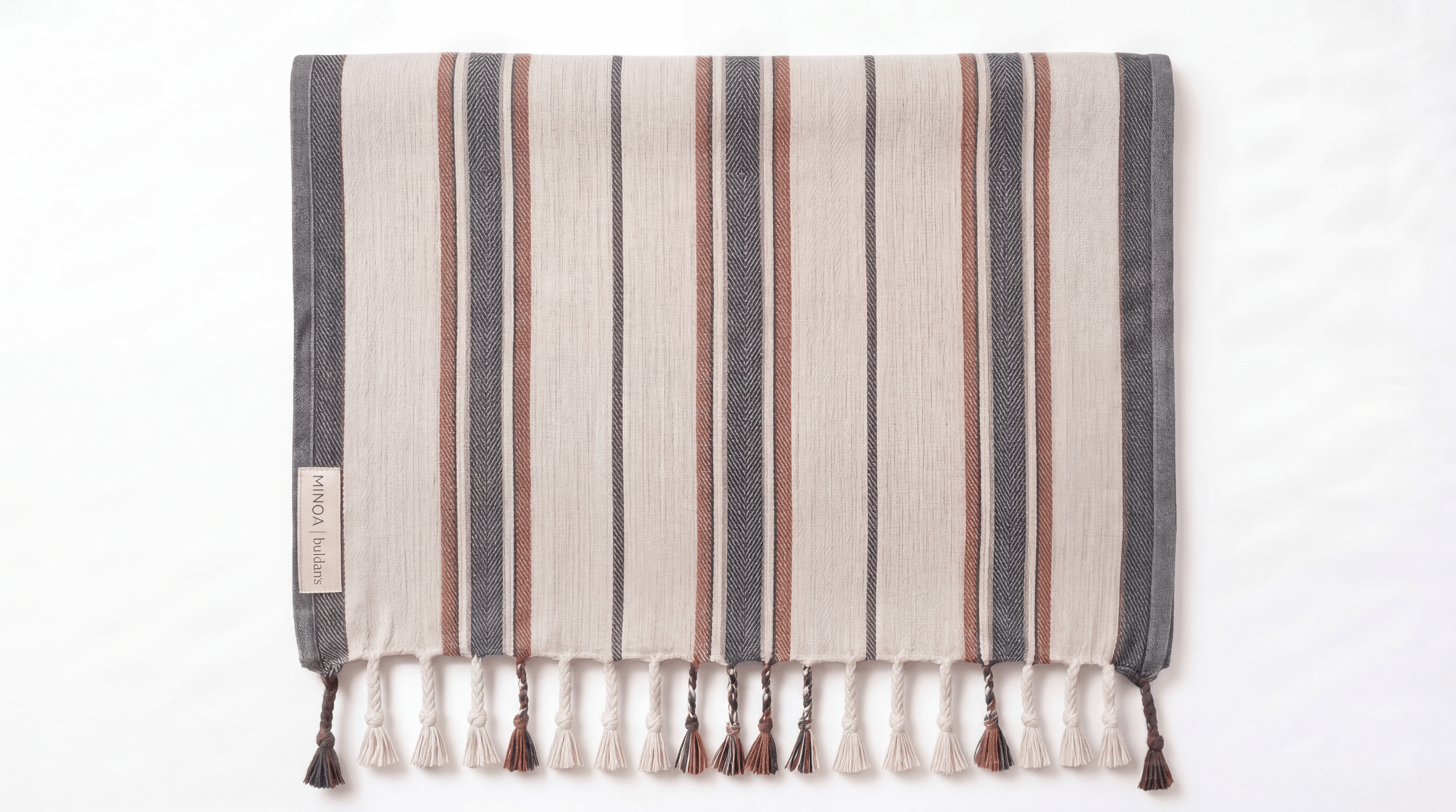 A rectangular woven towel with vertical stripes in beige, gray, and rust brown, finished with fringe tassels along the bottom edge. A small fabric label is stitched on the left side.
