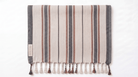 A rectangular woven towel with vertical stripes in beige, gray, and rust brown, finished with fringe tassels along the bottom edge. A small fabric label is stitched on the left side.