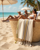 A woven straw tote bag on a sandy beach with a cream cotton towel draped over the side. The towel has green, blue, and red stripes with white tassels.