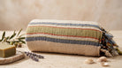 A striped cotton Turkish towel with fringed tassels, styled on a stone surface with a bar of soap, lavender sprigs, and seashells against a neutral beige background.