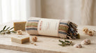 A rolled towel with fringe and a cream paper band reading MINOA, styled on a stone surface with soap bars, dried flowers, and shells in a soft neutral setting.