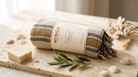 A rolled cotton towel with a cream paper band label reading MINOA, styled on a stone surface with soap, shells, dried flowers, and greenery.