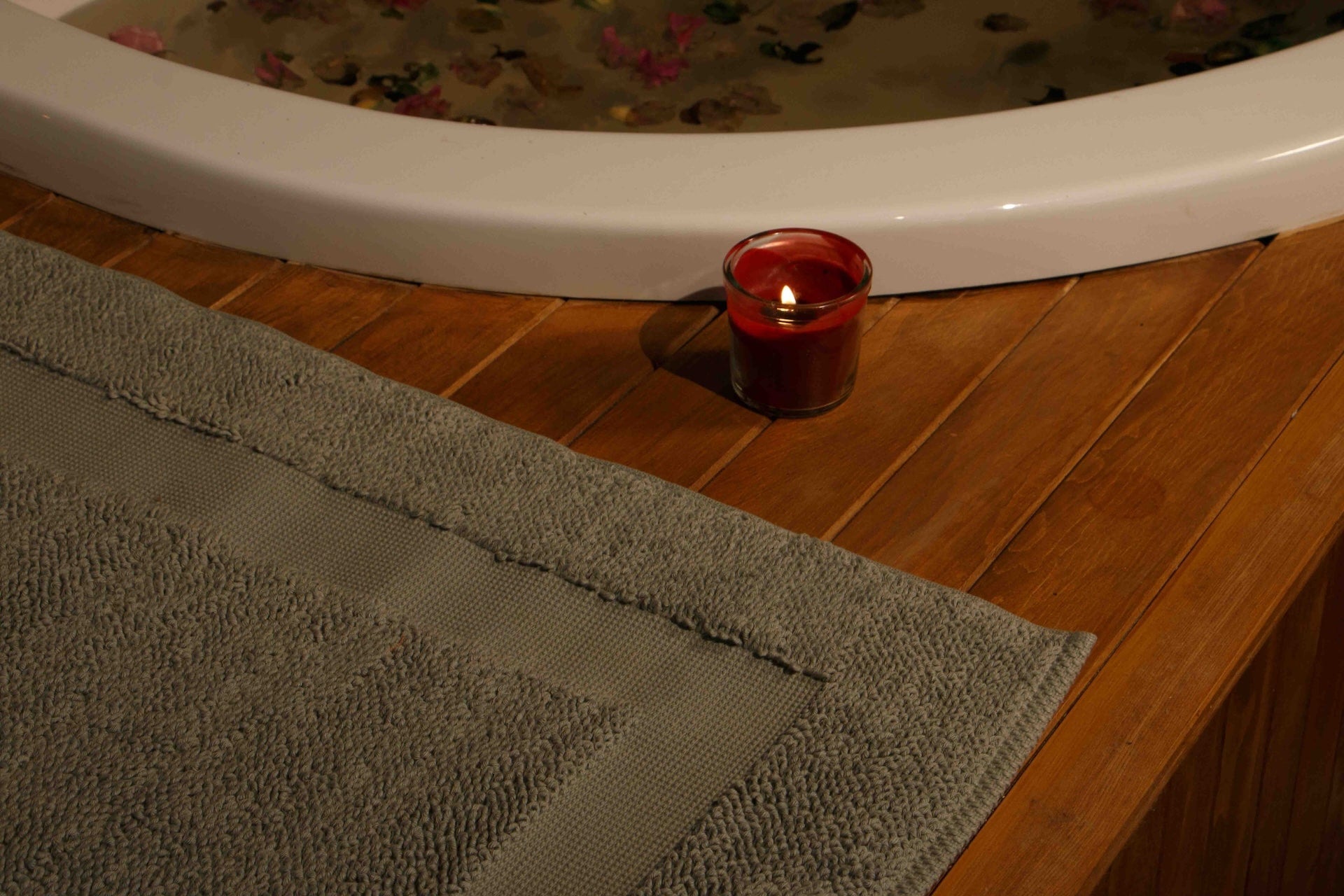 Candle in a red holder on a wooden surface next to a bathtub with a Minoa's Kaia Mat showcasing spa feeling 