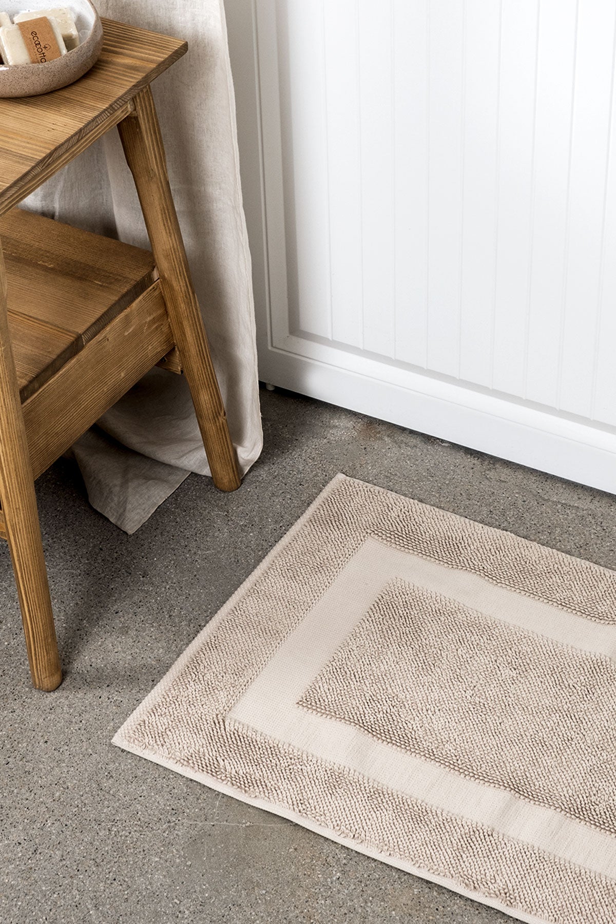 Kaia Aegean Cotton Bath Mat in Whispering Sands, placed in front of a bathroom door, showcasing its soft texture, warm hue, and luxurious quality
