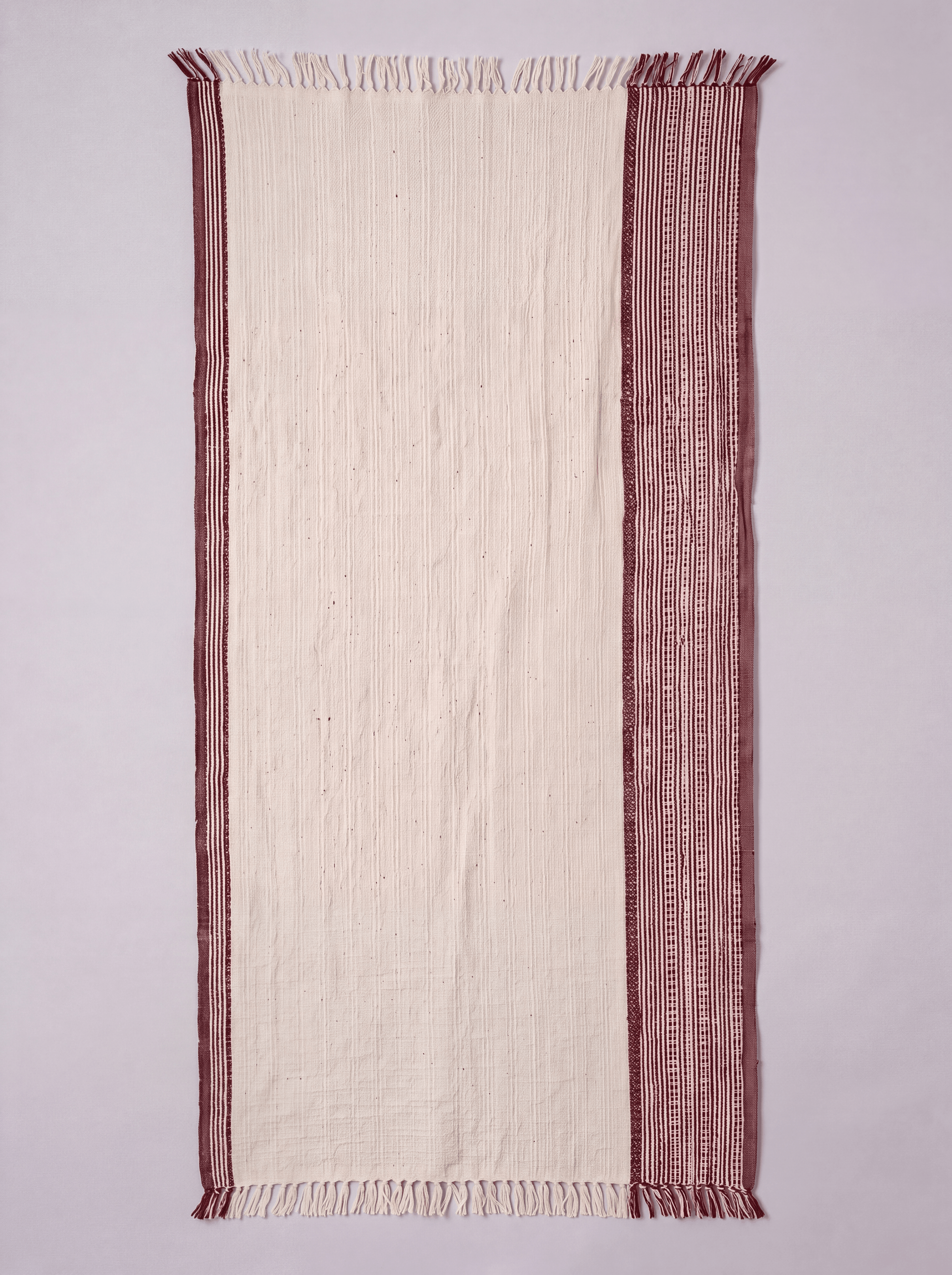 A rectangular cream-colored woven towel with fringed edges and a wide dusty rose patterned border, laid flat against a light gray background.