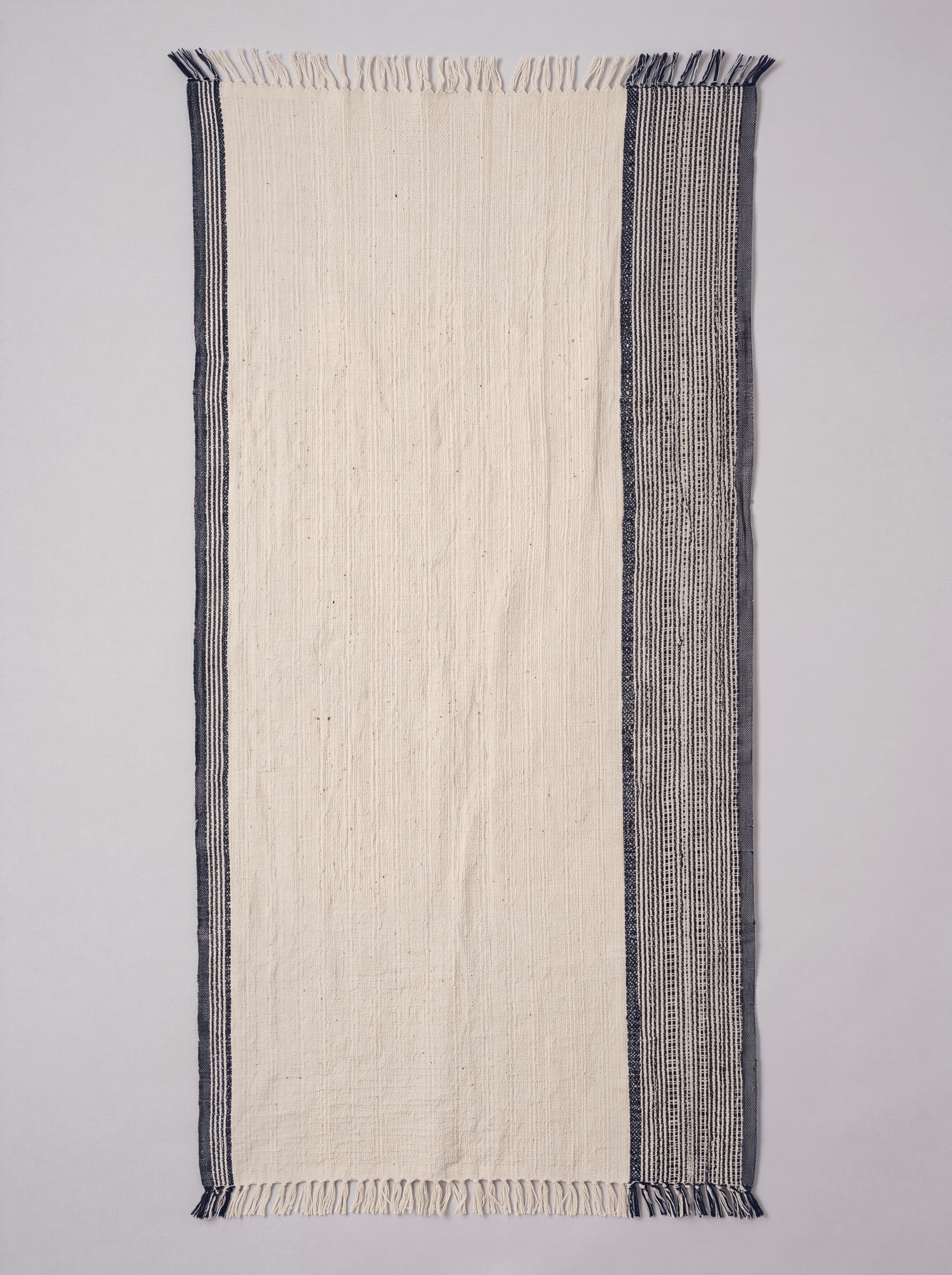 A rectangular woven towel featuring a wide off-white center panel, thin dark navy borders on the left, and a wider dark blue vertical patterned band on the right, finished with short fringe on both ends.