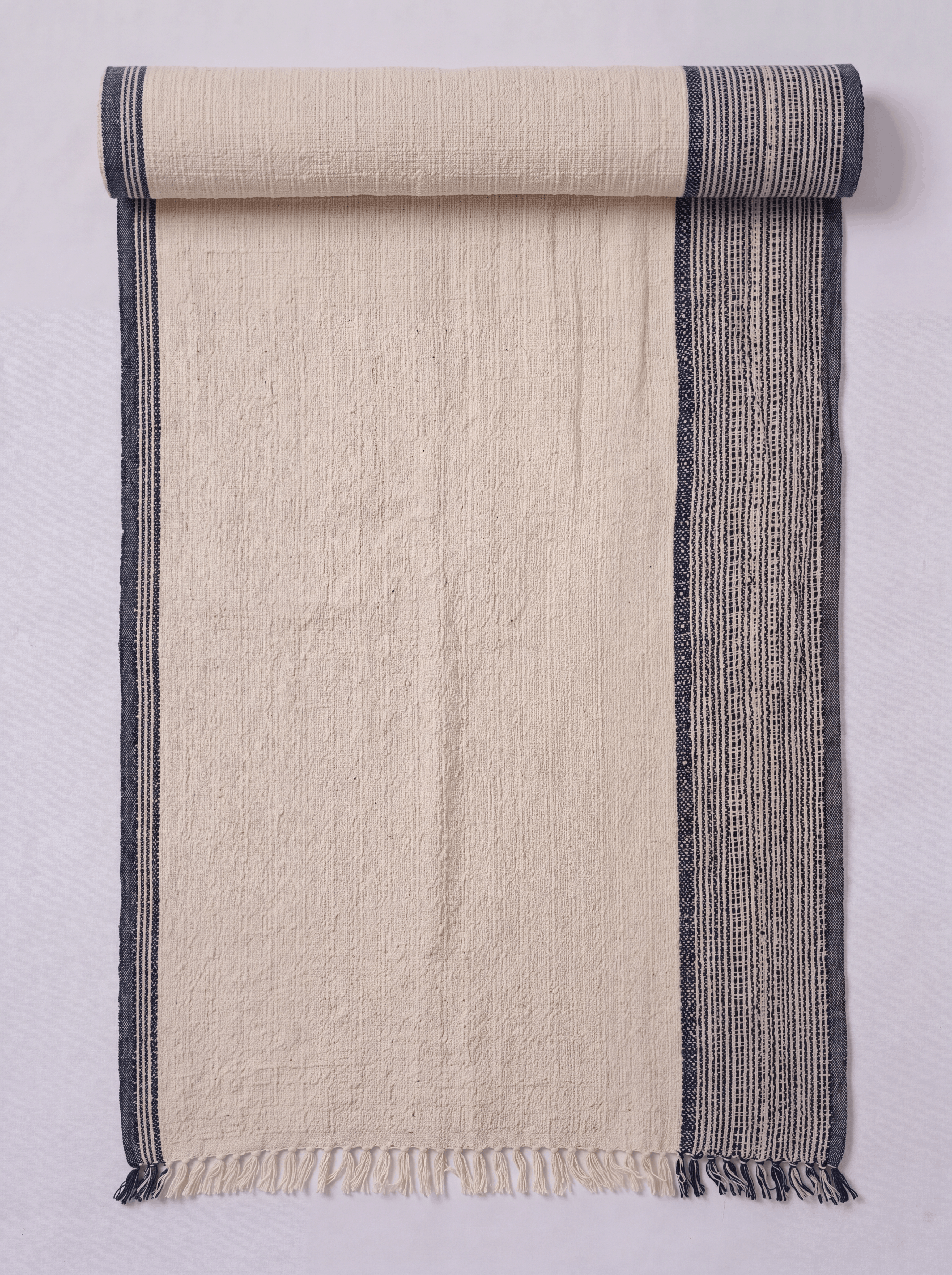 A cream-colored woven towel shown partially rolled at the top. It features a textured off-white center with a dark gray patterned border on the right side and a solid dark gray stripe on the left edge, finished with a fringed hem.
