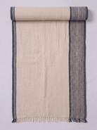 A cream-colored woven towel shown partially rolled at the top. It features a textured off-white center with a dark gray patterned border on the right side and a solid dark gray stripe on the left edge, finished with a fringed hem.