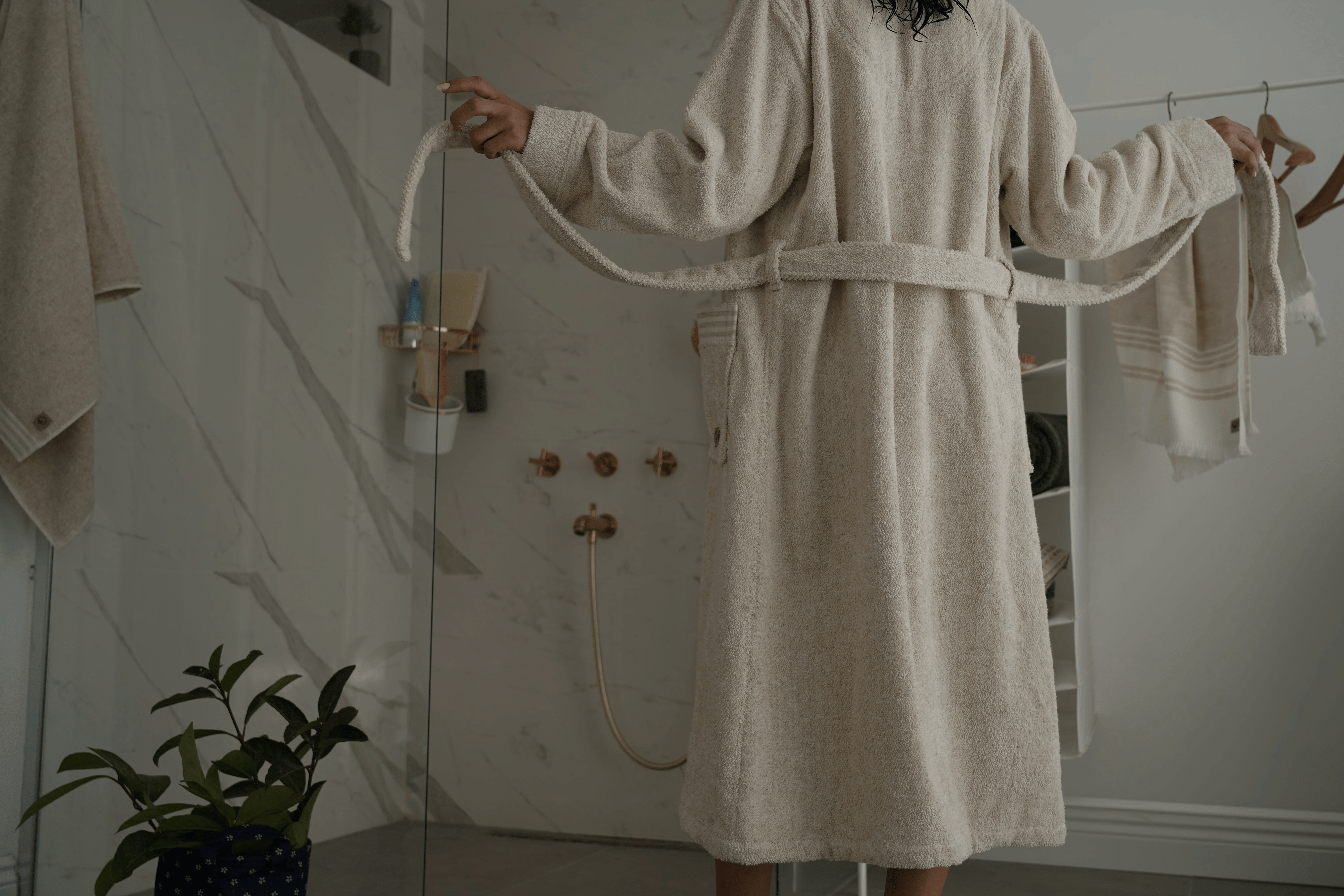 Person wearing Minoa's luxury bath robe in a bathroom setting with a plant and shower visible