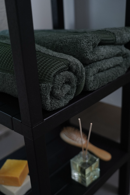 Green towels on a black shelf with a wooden brush and glass container in the background.