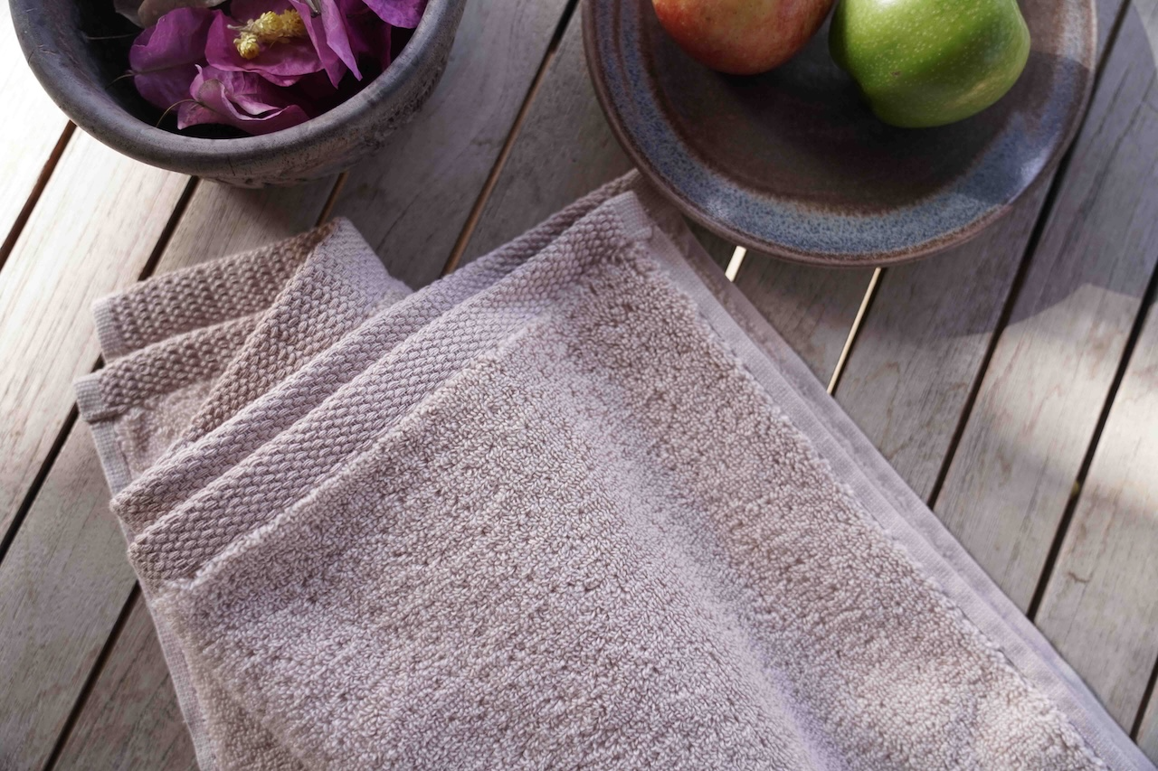 Stack of gray towels on a wooden surface with a bowl of flowers and apples in the background.