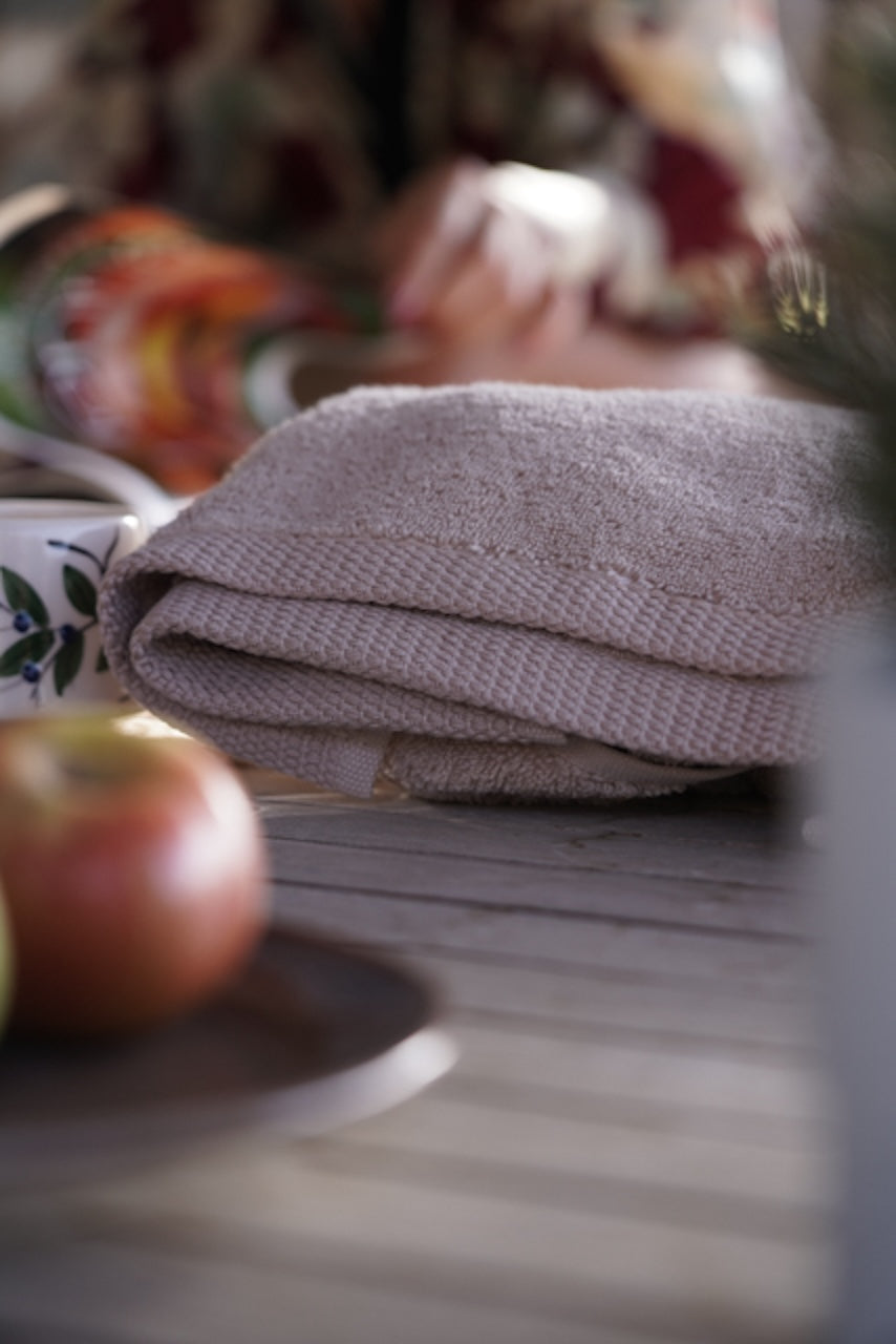 Folded beige towel on a wooden surface with an apple and a cup in the foreground.
