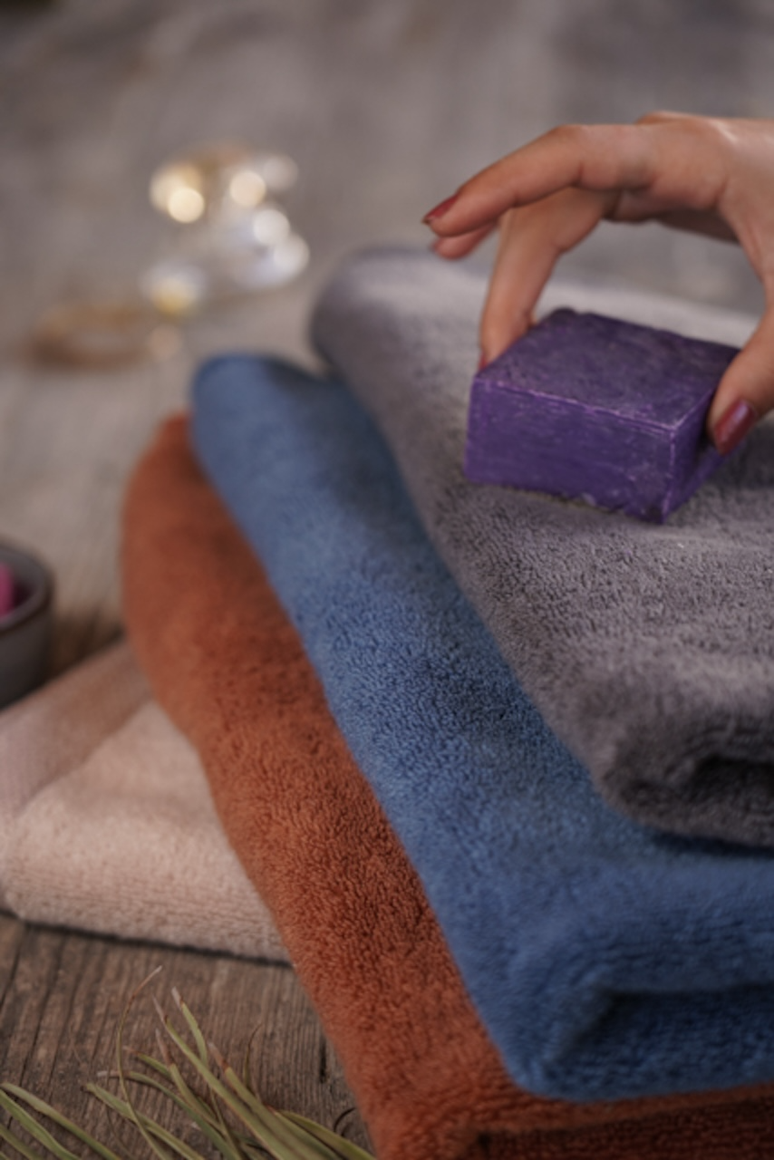 Hand holding a purple bar of soap over stacked colorful towels with a blurred background.