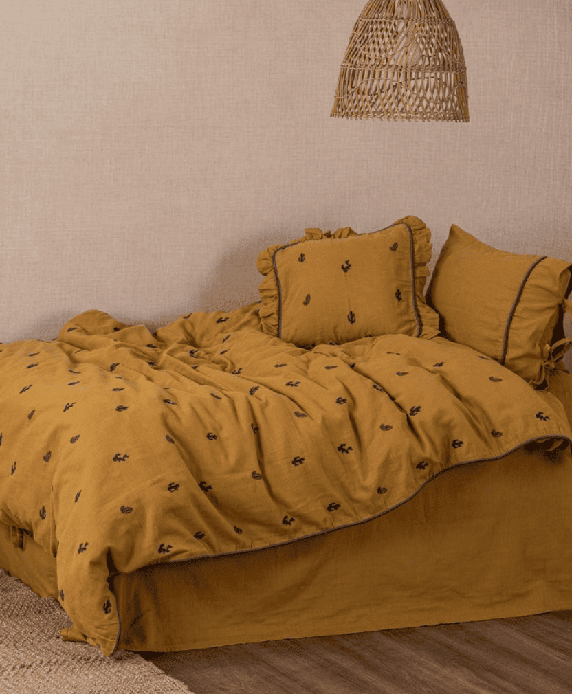 A beige/mustard colored bedding set with a floral pattern displayed on a bed in a room with a wicker lampshade.