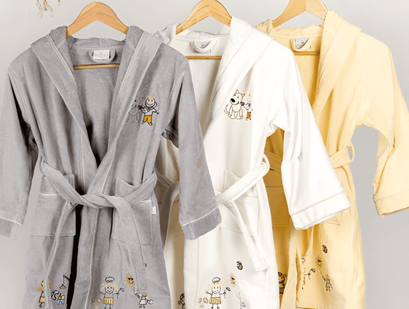 Three bathrobes in gray, white, and yellow hanging on a rack against a light gray background.