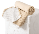 Two folded blankets, one white with fringes and the other beige, on a light wooden surface.