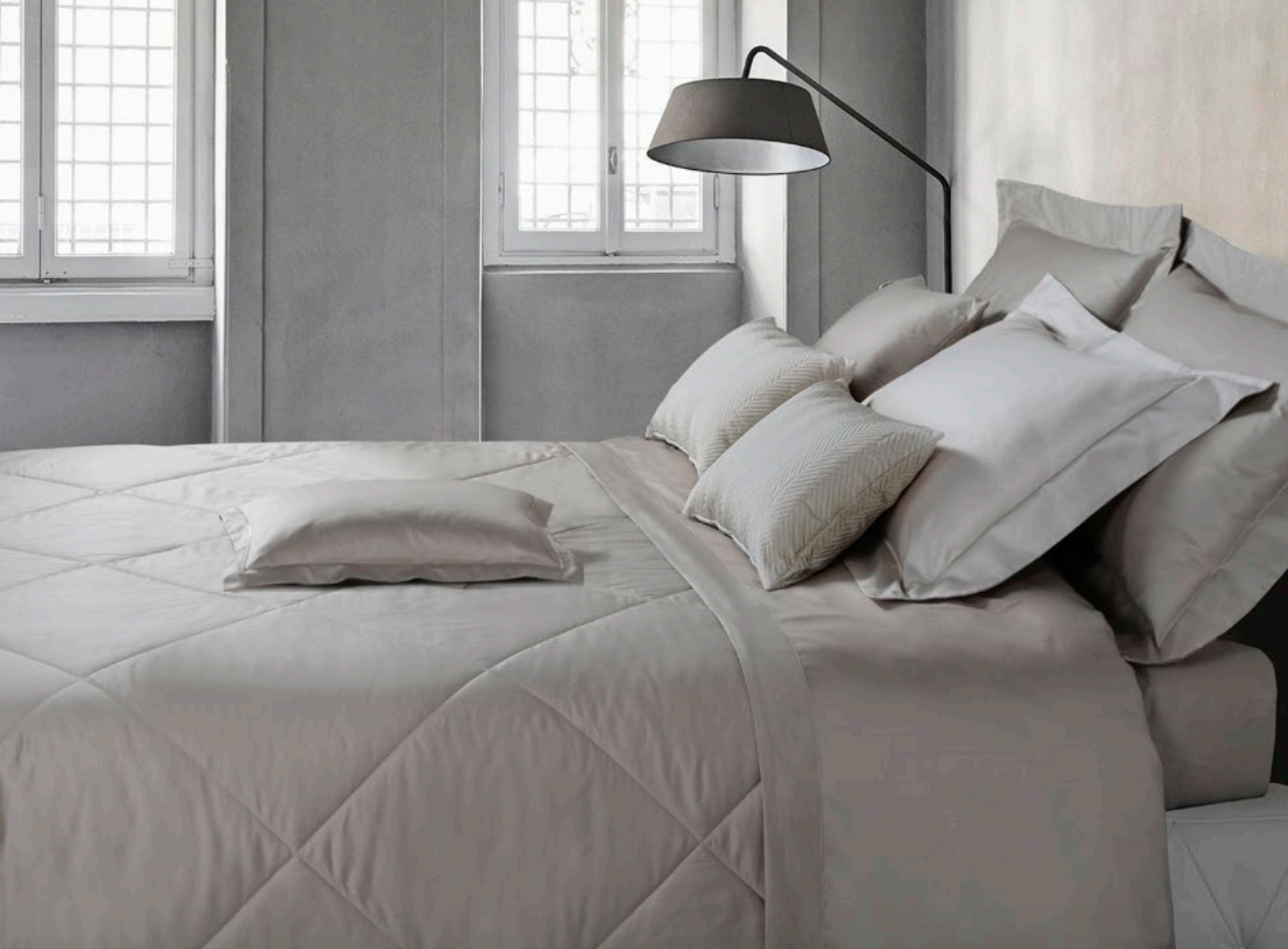 Minimalist bedroom with soft grey bedding, layered pillows, and natural light from two windows.