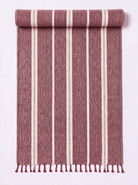 A rolled and unrolled woven towel featuring vertical stripes in dusty rose, cream, and white. The fabric has a textured ikat-like weave and finished with a fringed tassel edge at the bottom, displayed against a clean white background.