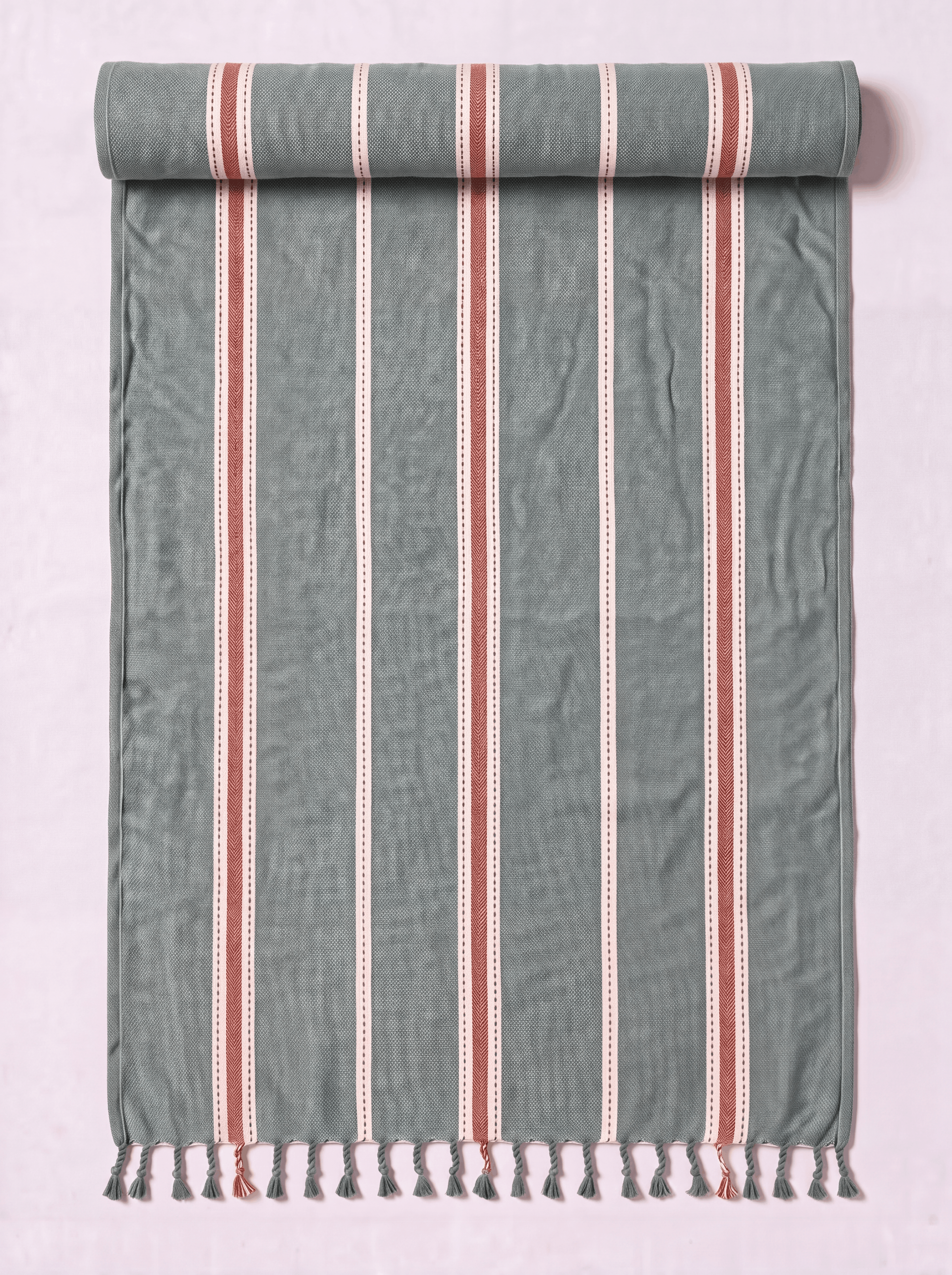 A rolled-out cotton Turkish towel with vertical stripes in sage green, terracotta red, and white, finished with a fringed tassel edge, shown against a light pink background.