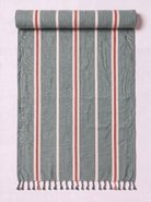 A rolled-out cotton Turkish towel with vertical stripes in sage green, terracotta red, and white, finished with a fringed tassel edge, shown against a light pink background.