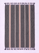 A rectangular woven towel with dark gray base, vertical stripes in beige, white, and muted pink, and dark purple fringe tassels on the top and bottom, laid flat on a light lavender background.