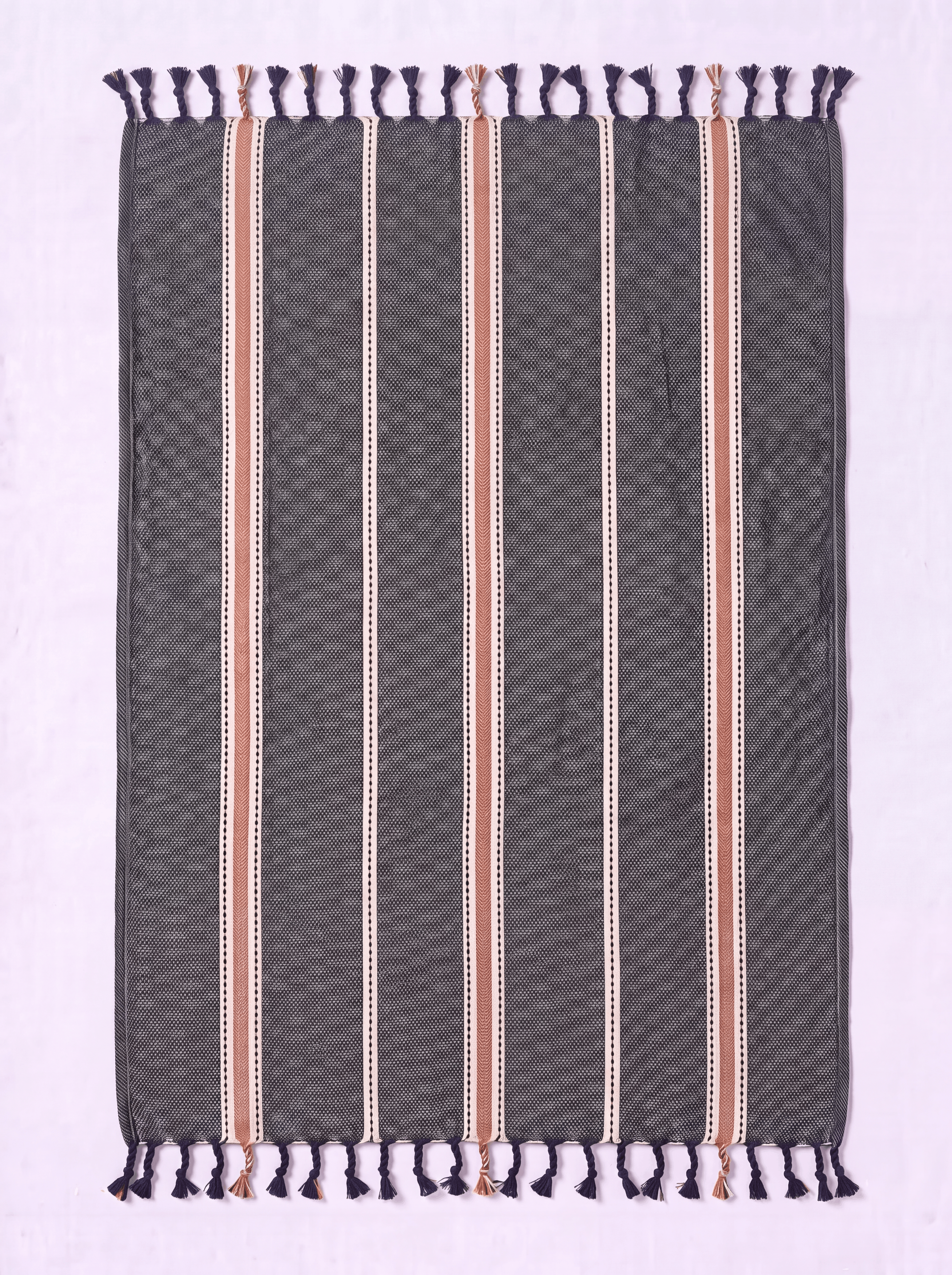 A rectangular woven towel with dark gray base, vertical stripes in beige, white, and muted pink, and dark purple fringe tassels on the top and bottom, laid flat on a light lavender background.