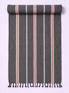 A dark gray woven towel with vertical stripes in cream, terracotta, and white, finished with fringe tassels at the bottom, shown partially rolled on a light lavender background.