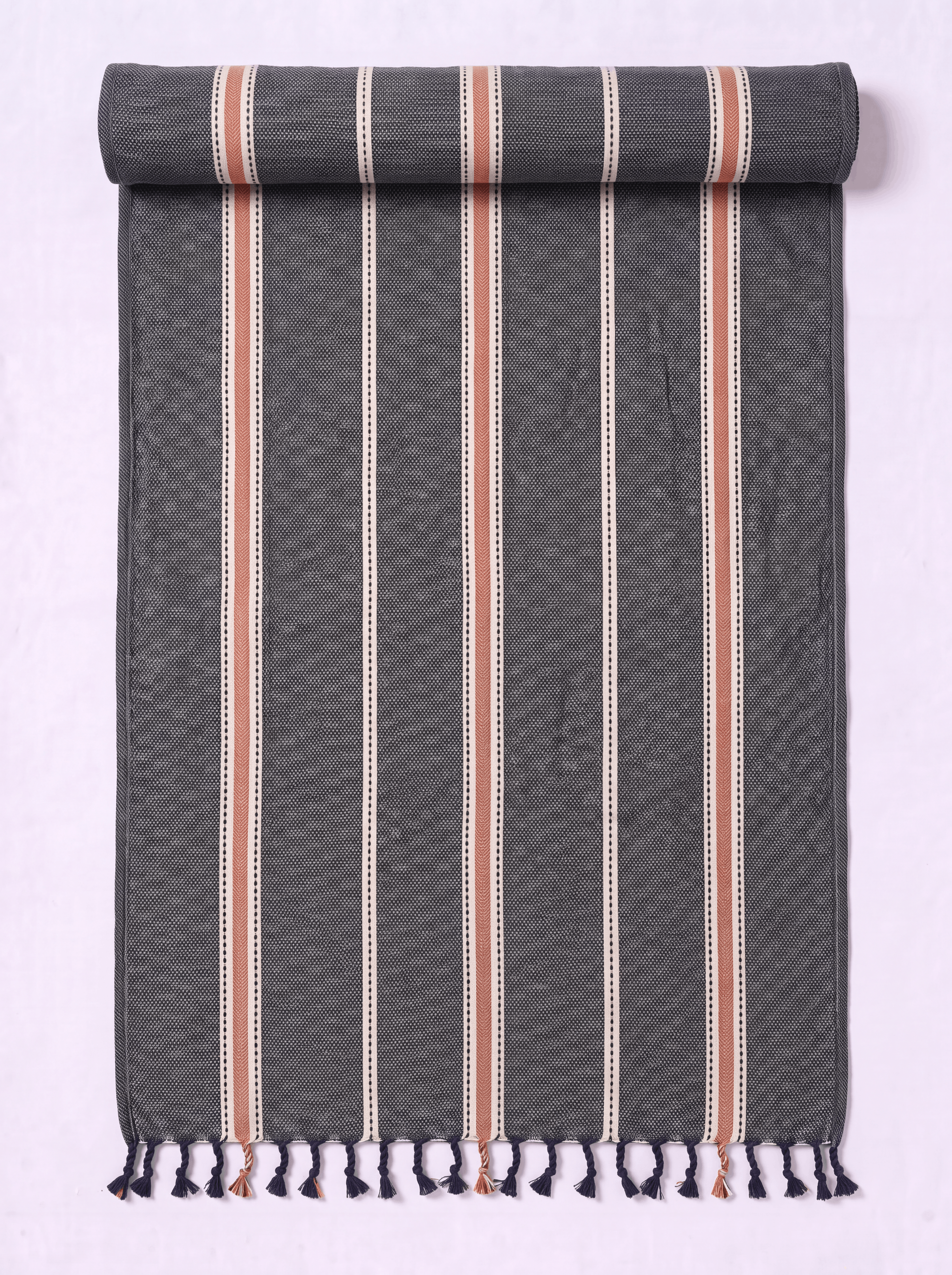 A dark gray woven towel with vertical stripes in cream, terracotta, and white, finished with fringe tassels at the bottom, shown partially rolled on a light lavender background.