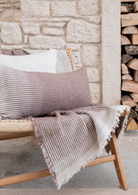 Minoa Amaya collection featuring a striped cushion and bed cover in soft earthy tones on a woven bench, elegant sustainable home decor with artisanal detail.