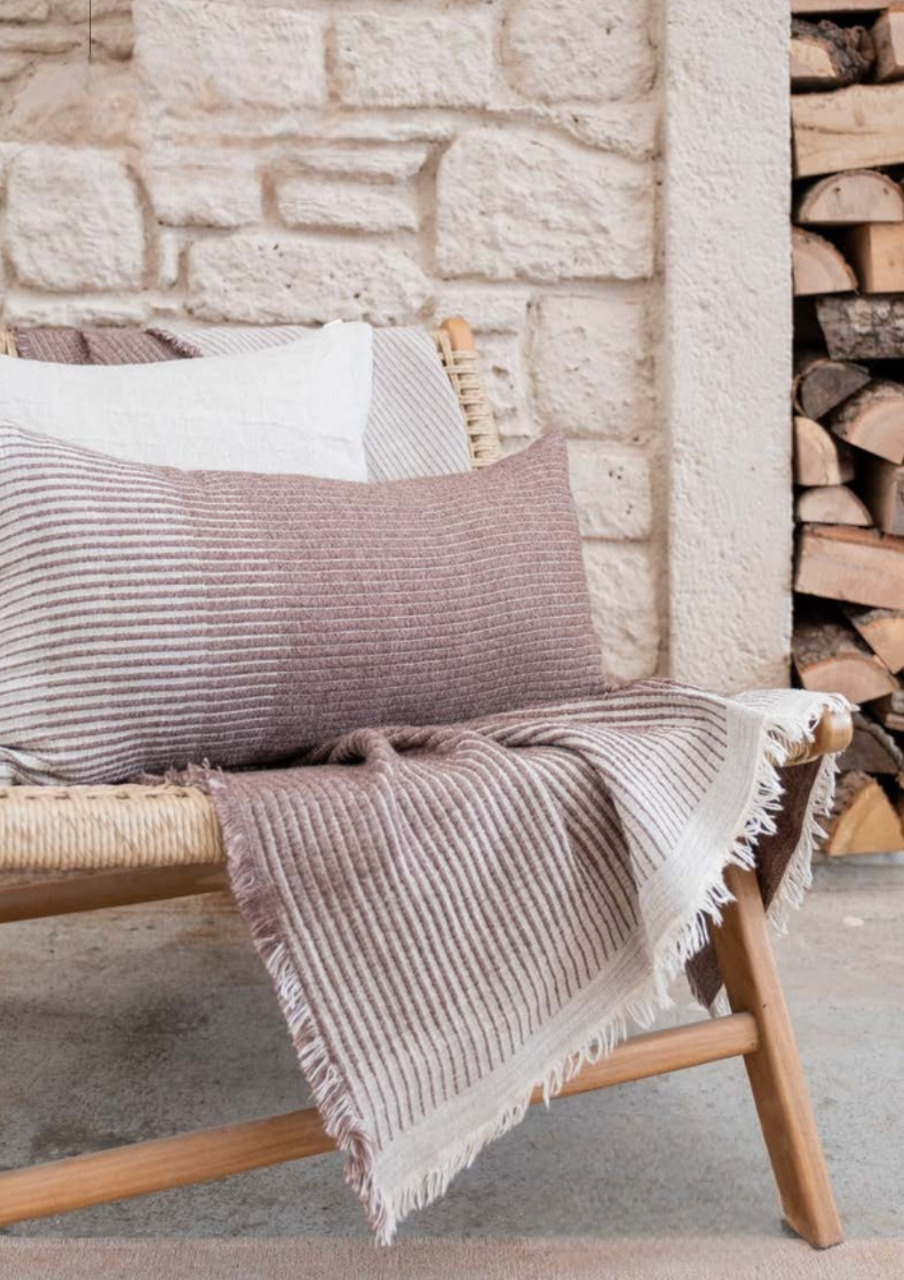 Minoa Amaya collection featuring a striped cushion and bed cover in soft earthy tones on a woven bench, elegant sustainable home decor with artisanal detail.