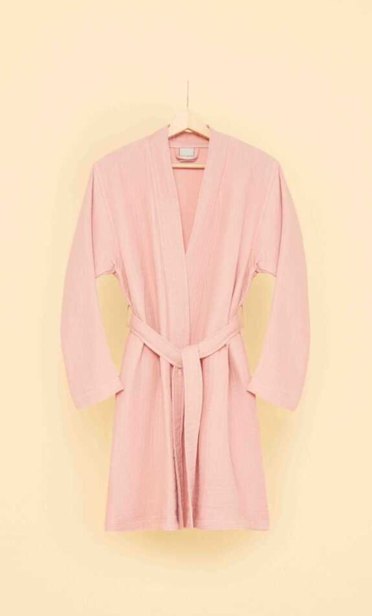 Blush pink bathrobe with belt tie hanging on wooden hanger against soft pastel background — minimalist eco-luxury loungewear aesthetic.