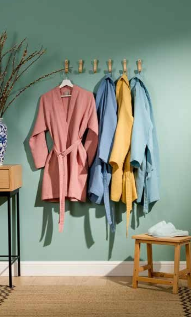 Four colorful bathrobes in pink, blue, yellow, and light blue hanging on wooden hooks against a green wall — trendy sustainable home essentials setup.