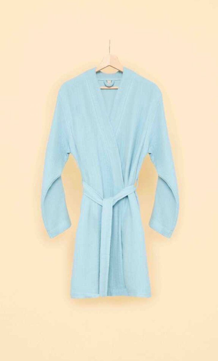 Light blue bathrobe with belt tie on wooden hanger against a soft beige background — minimalist sustainable loungewear aesthetic.